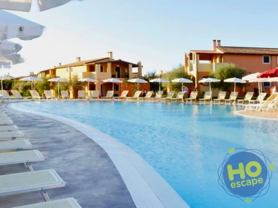 Piscina con lettini e ombrelloni bianchi circondata dagli edifici color arancio del resort