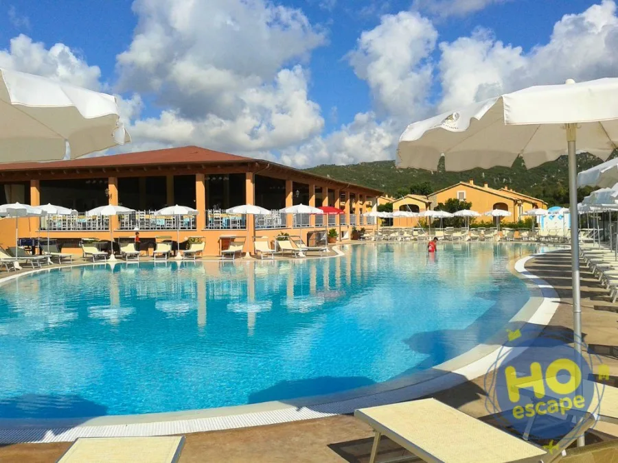 Piscina curva con lettini e ombrelloni bianchi, ristorante coperto e cielo nuvoloso