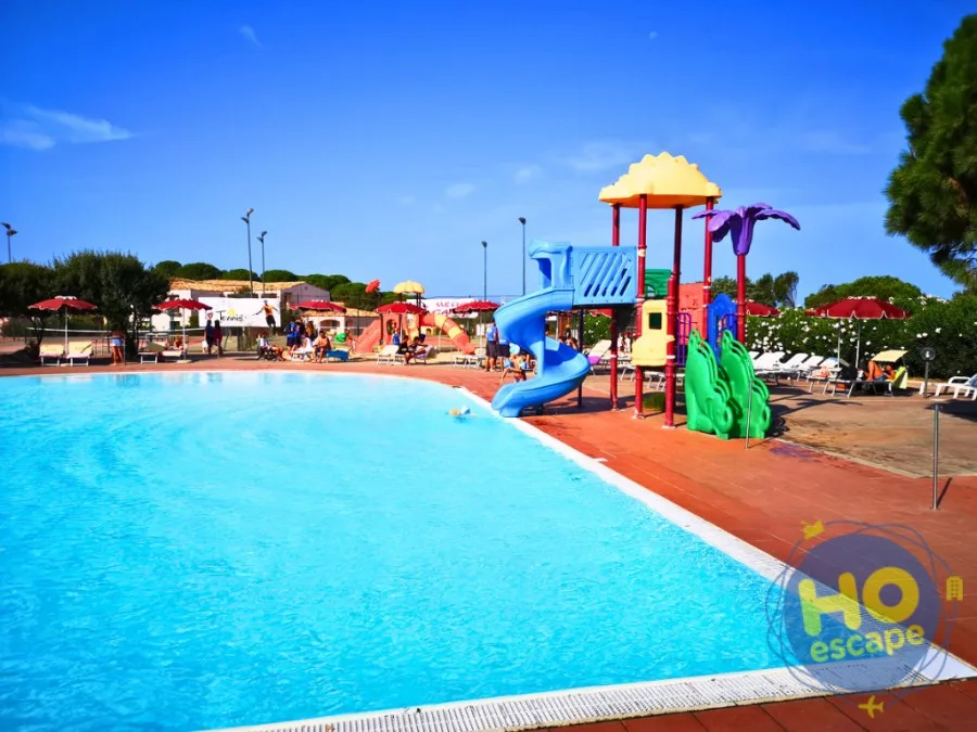 Piscina con area giochi colorata e scivolo per bambini circondata da ombrelloni