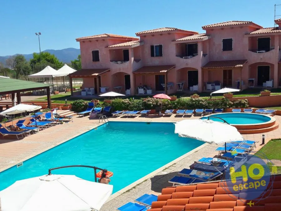 Vista panoramica della piscina del residence con lettini blu, ombrelloni e edifici rosa sullo sfondo delle montagne