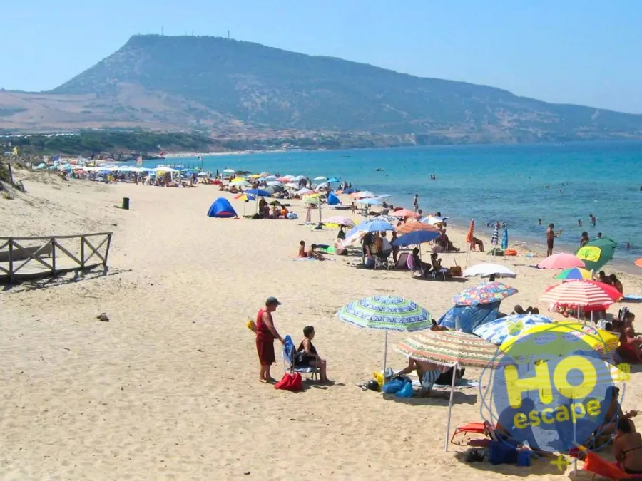 Spiaggia sabbiosa affollata di ombrelloni colorati con mare turchese e promontorio sullo sfondo