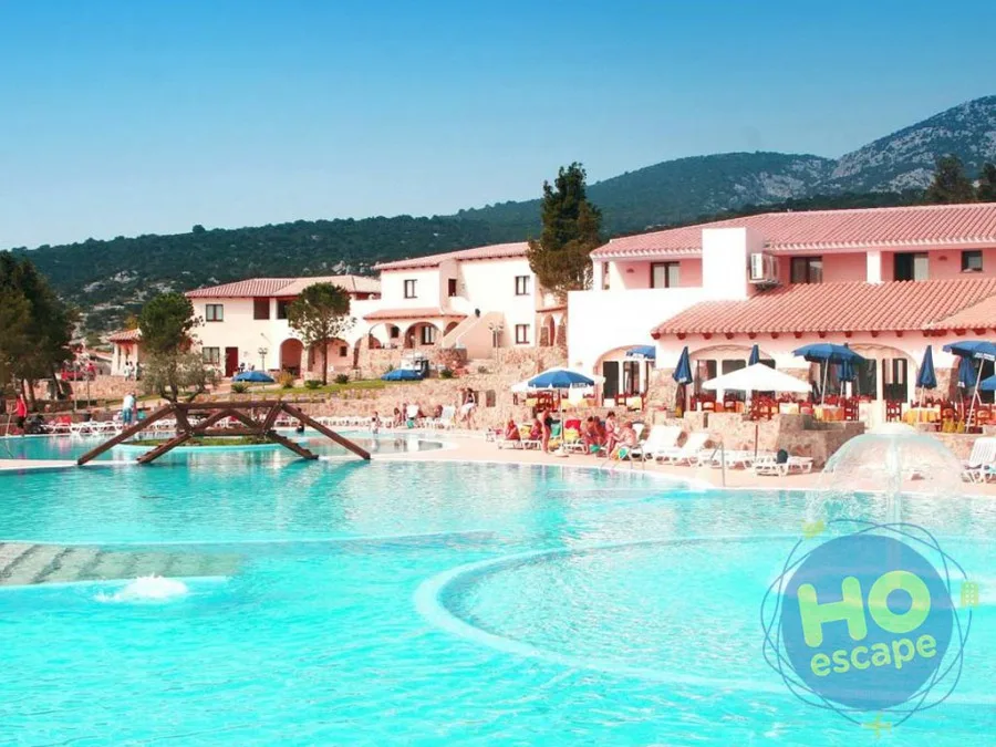 Ampia piscina con acqua turchese e fontane, circondata da lettini e ombrelloni con edifici dell'hotel e montagne sullo sfondo