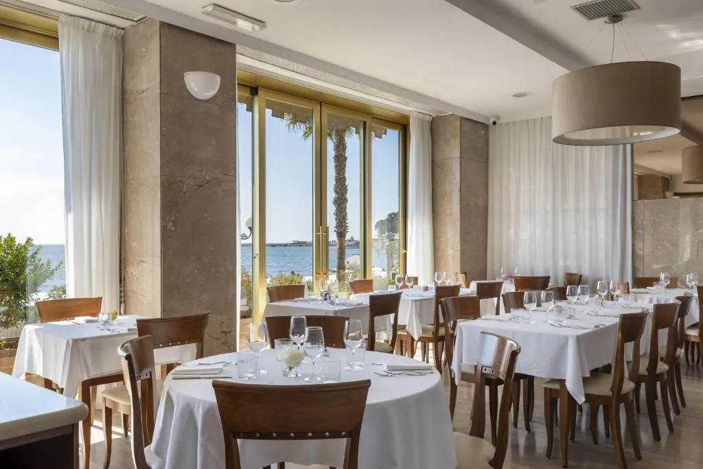 Sala ristorante con tavoli apparecchiati di bianco e ampie vetrate con vista sul mare e palma