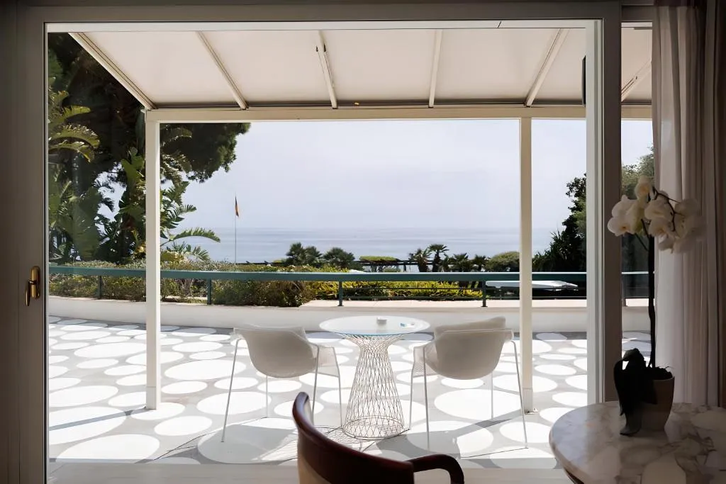 Terrazza luminosa con tavolino, sedie bianche e vista sul mare attraverso ampie vetrate