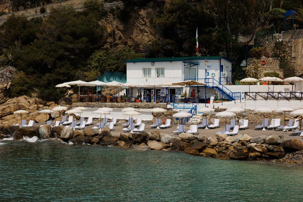 Stabilimento balneare con lettini bianchi e ombrelloni su spiaggia di ciottoli ai piedi della scogliera