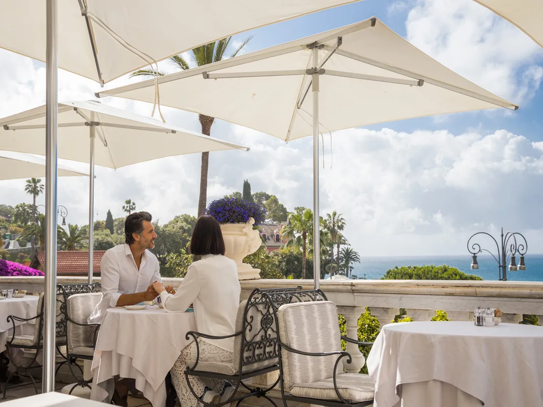 Coppia seduta in terrazza sotto ombrelloni bianchi con vista sul mare e giardini