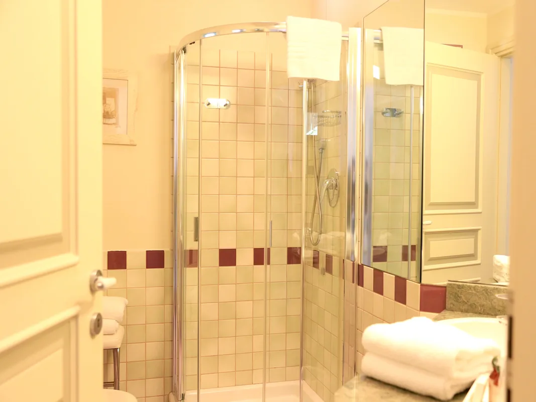 Bagno con doccia angolare in vetro, piastrelle beige e asciugamani bianchi