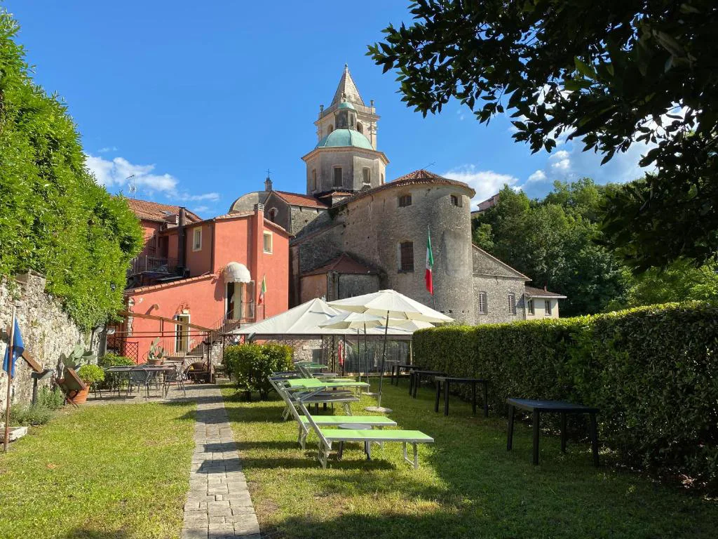 Esterno dell'hotel con giardino, ombrelloni e campanile storico sotto un cielo azzurro