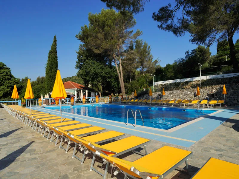 Piscina all'aperto con file di lettini e ombrelloni gialli sotto i pini