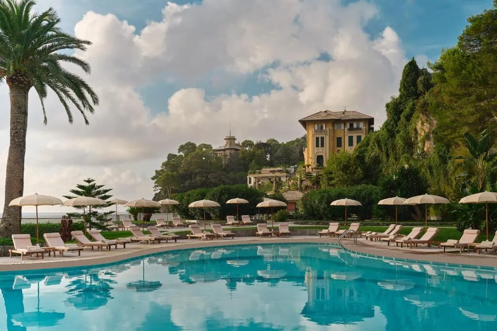 Piscina dell'hotel con lettini e ombrelloni bianchi, palme e villa storica sullo sfondo al tramonto