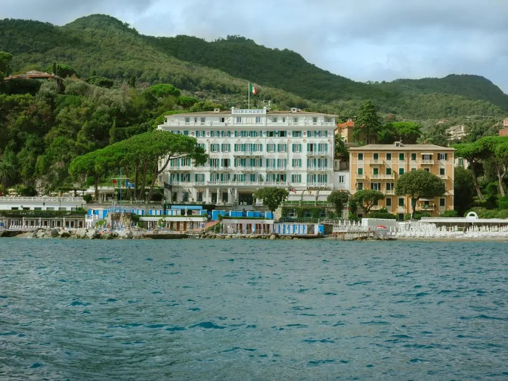 Vista dal mare dell'hotel storico bianco con spiaggia attrezzata, immerso nella vegetazione collinare