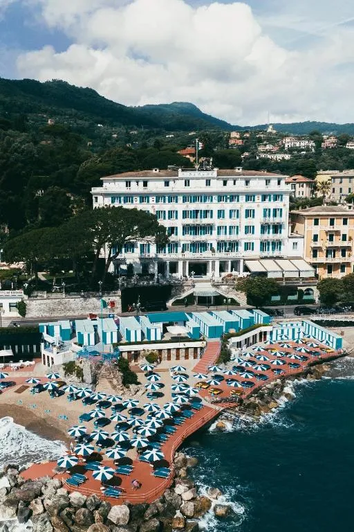 Vista aerea dell'elegante hotel storico bianco fronte mare con spiaggia attrezzata e ombrelloni a strisce blu