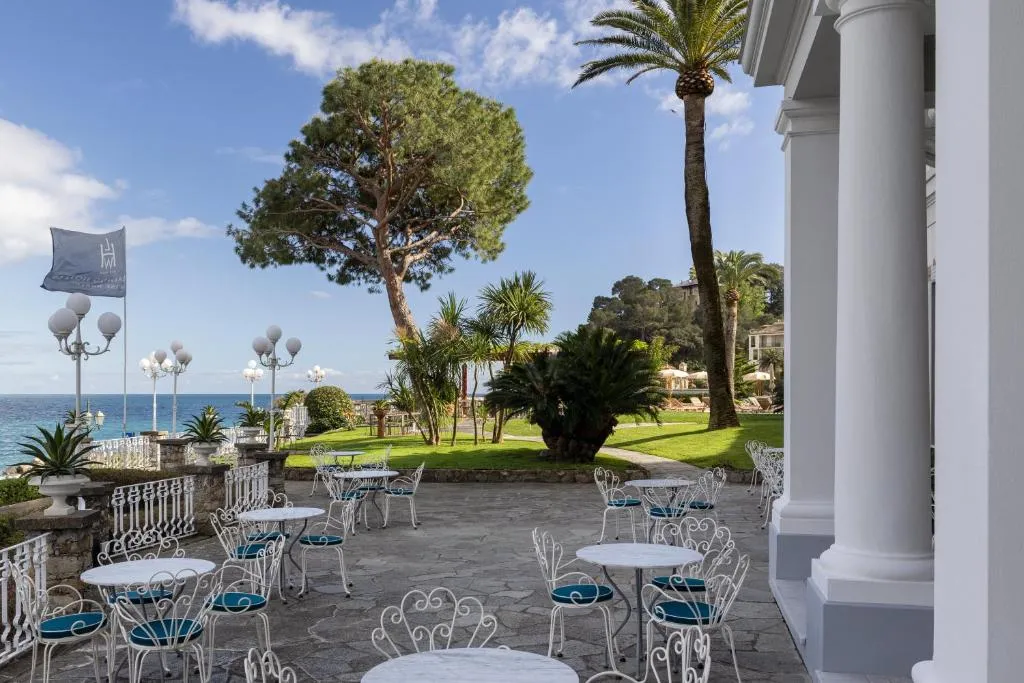 Terrazza dell'hotel con tavolini e sedie in ferro battuto, palme e giardino curato con vista sul mare