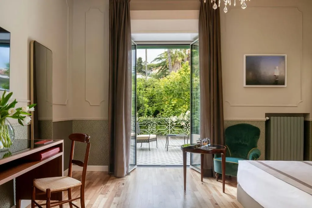 Elegante camera con scrivania in legno, poltrona verde e ampia porta finestra che si apre su una terrazza con giardino