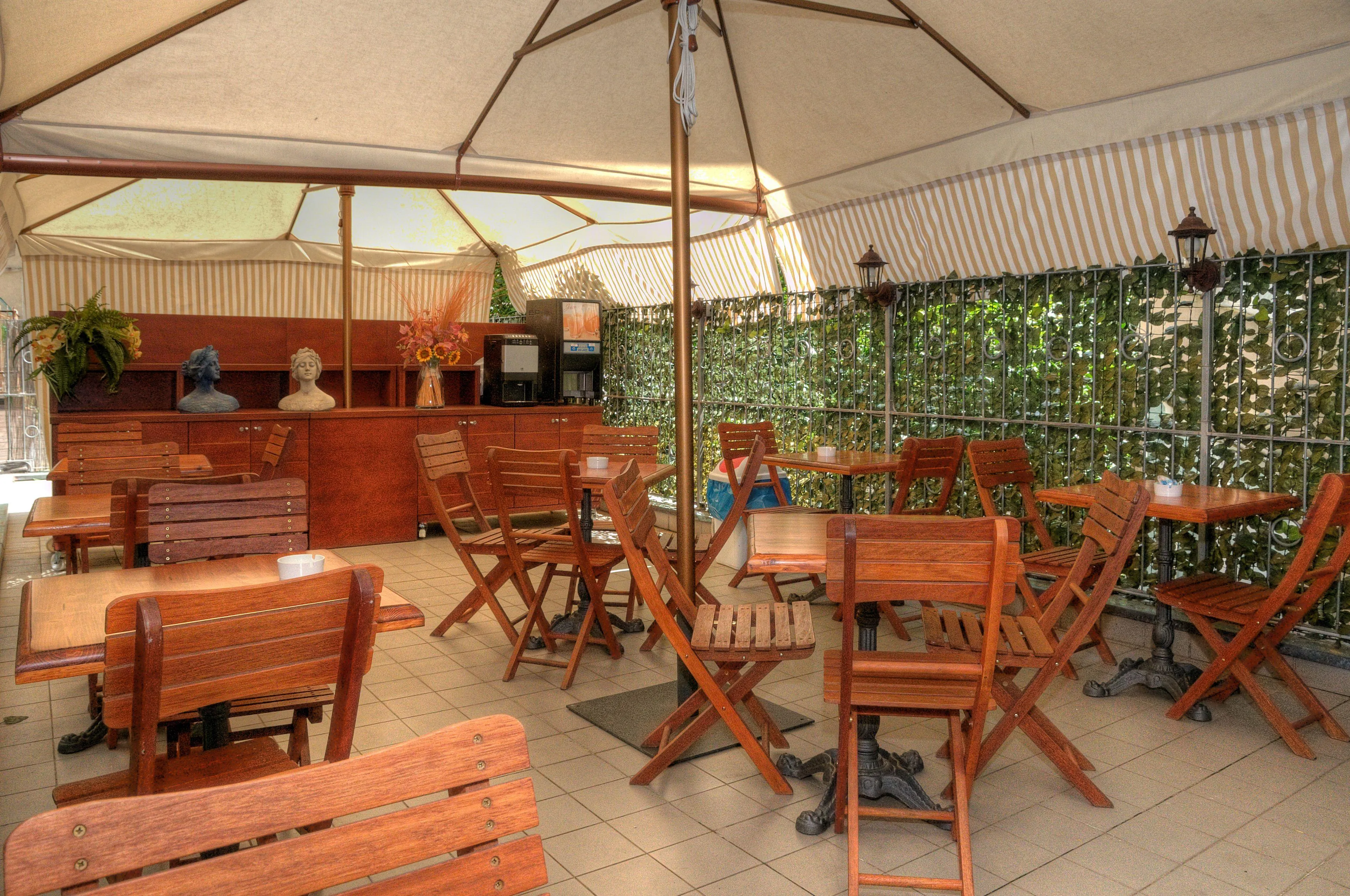 Terrazza esterna coperta con ombrellone, tavoli e sedie in legno e siepe verde