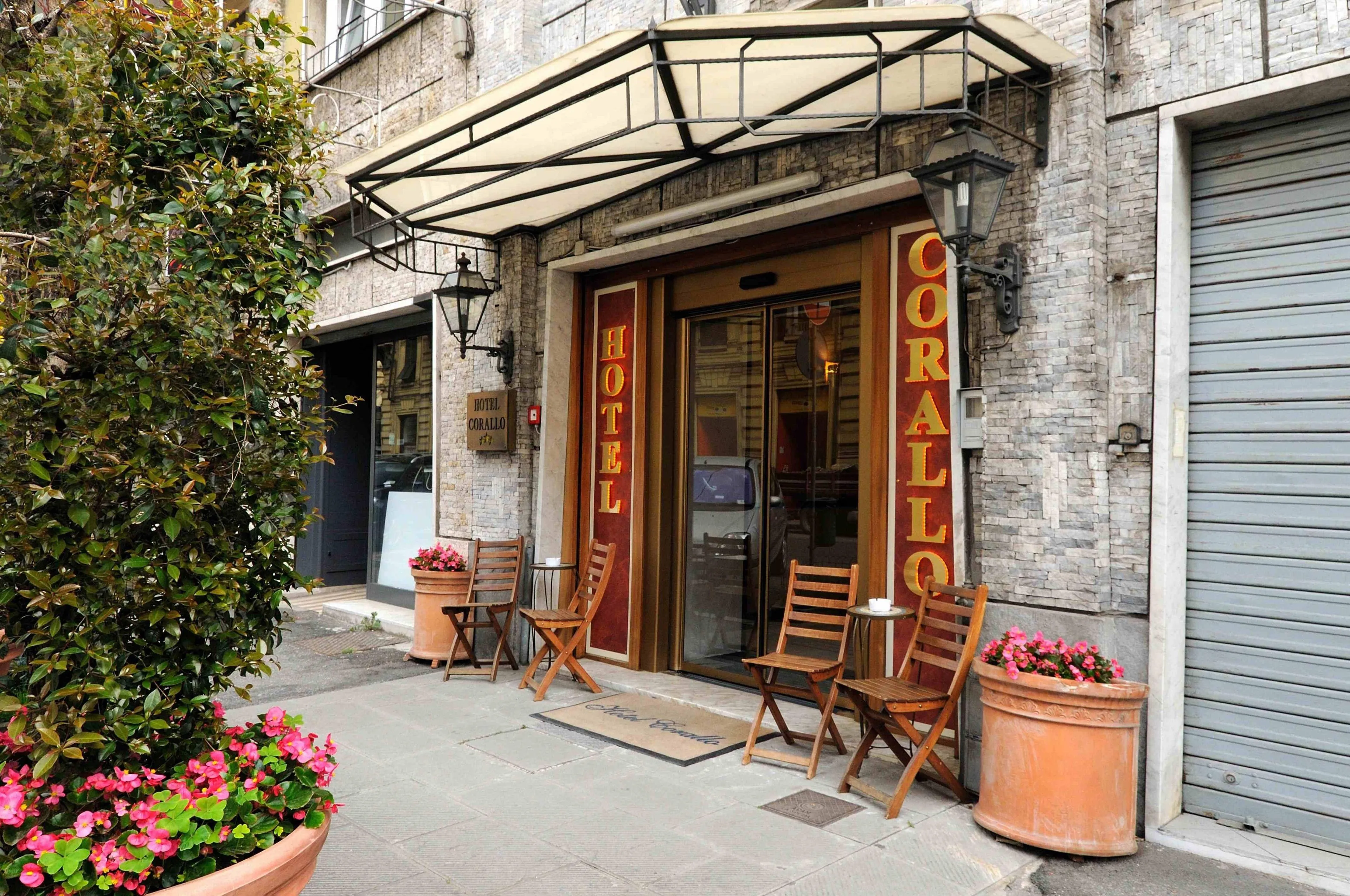 Ingresso dell'Hotel Corallo con insegna rossa e dorata, tenda, sedie in legno e piante fiorite