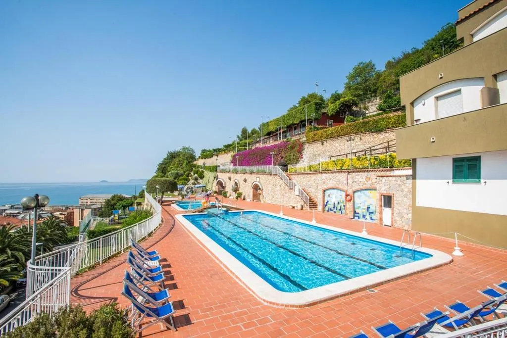 Piscina su terrazza panoramica con vista mare, lettini blu, fioriture colorate e edifici del residence