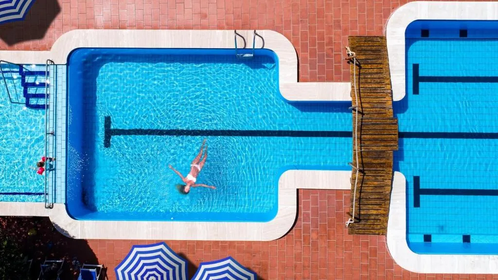Vista aerea di una piscina con persona che nuota, ponticello in legno e ombrelloni a righe blu