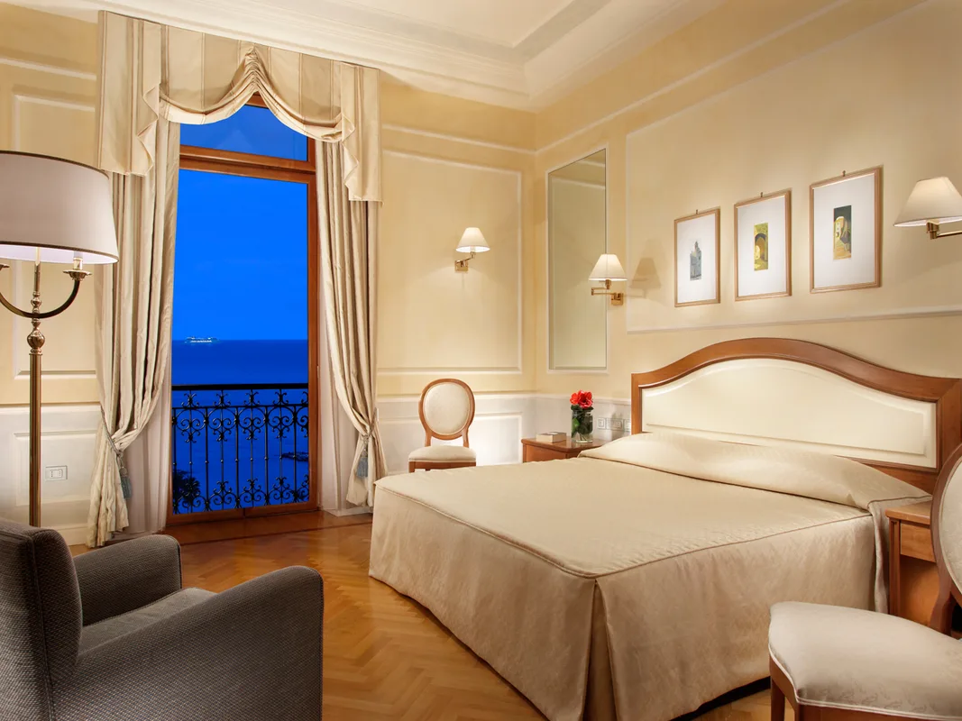 Camera classica con tonalità calde, arredi in legno e balcone con vista serale sul mare