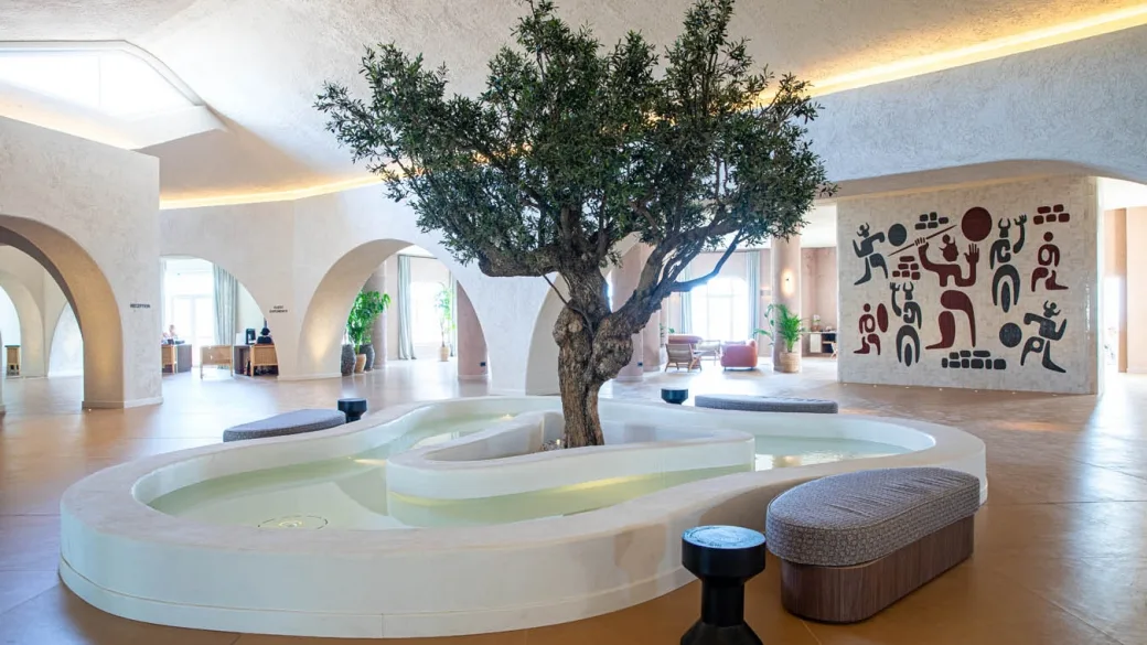 Hall dell'hotel con ulivo centrale, fontana bianca e murale decorativo su pareti chiare