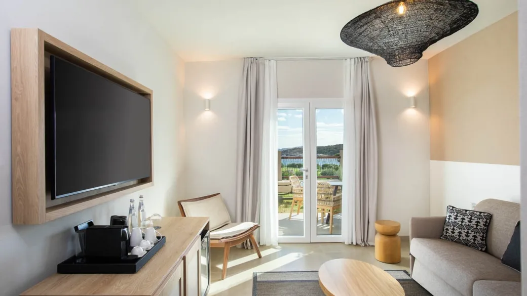 Soggiorno di camera con TV a parete, divano, poltrona e portafinestra con vista mare