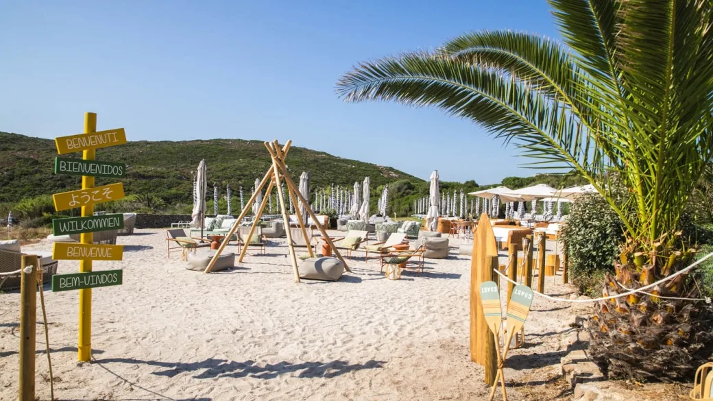 Beach club sulla sabbia con sedute lounge, ombrelloni e cartello di benvenuto multilingue
