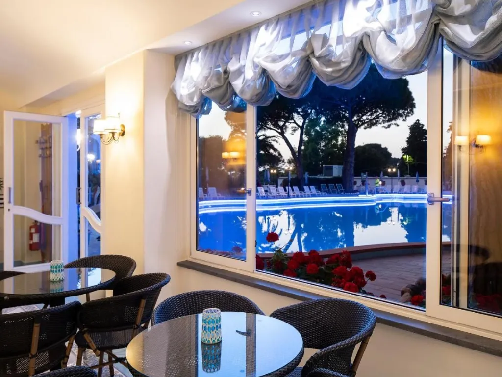 Sala interna con tavolini e vista sulla piscina illuminata attraverso ampie vetrate